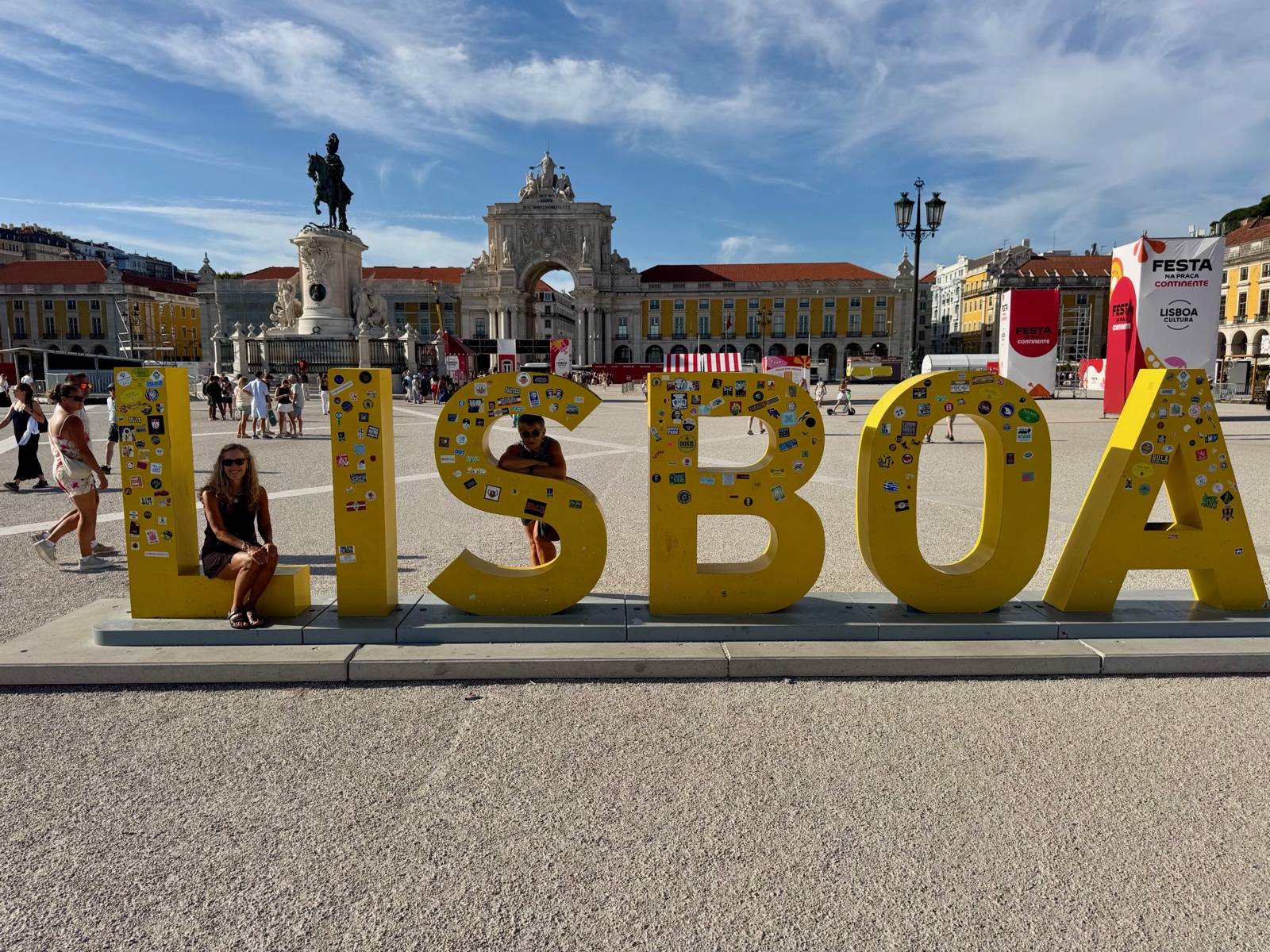 Giant yellow letters spelling LISBOA, covered in stickers, in a wide sunlit plaza in Lisbon.
          Two people pose among the letters. Behind them stand an equestrian statue, a large stone arch,
          and yellow buildings under a blue sky.