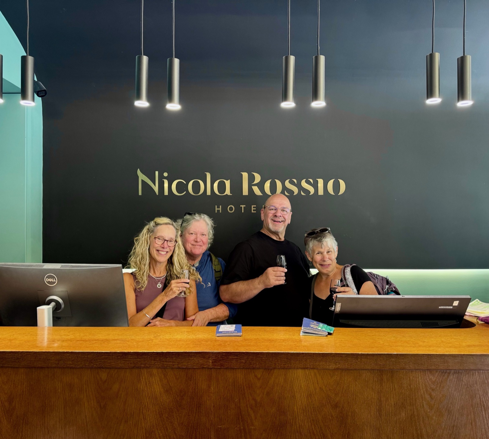 Four smiling travelers standing behind a hotel reception desk beneath a sign reading
          Nicola Rossio Hotel, each raising a small glass in a toast.