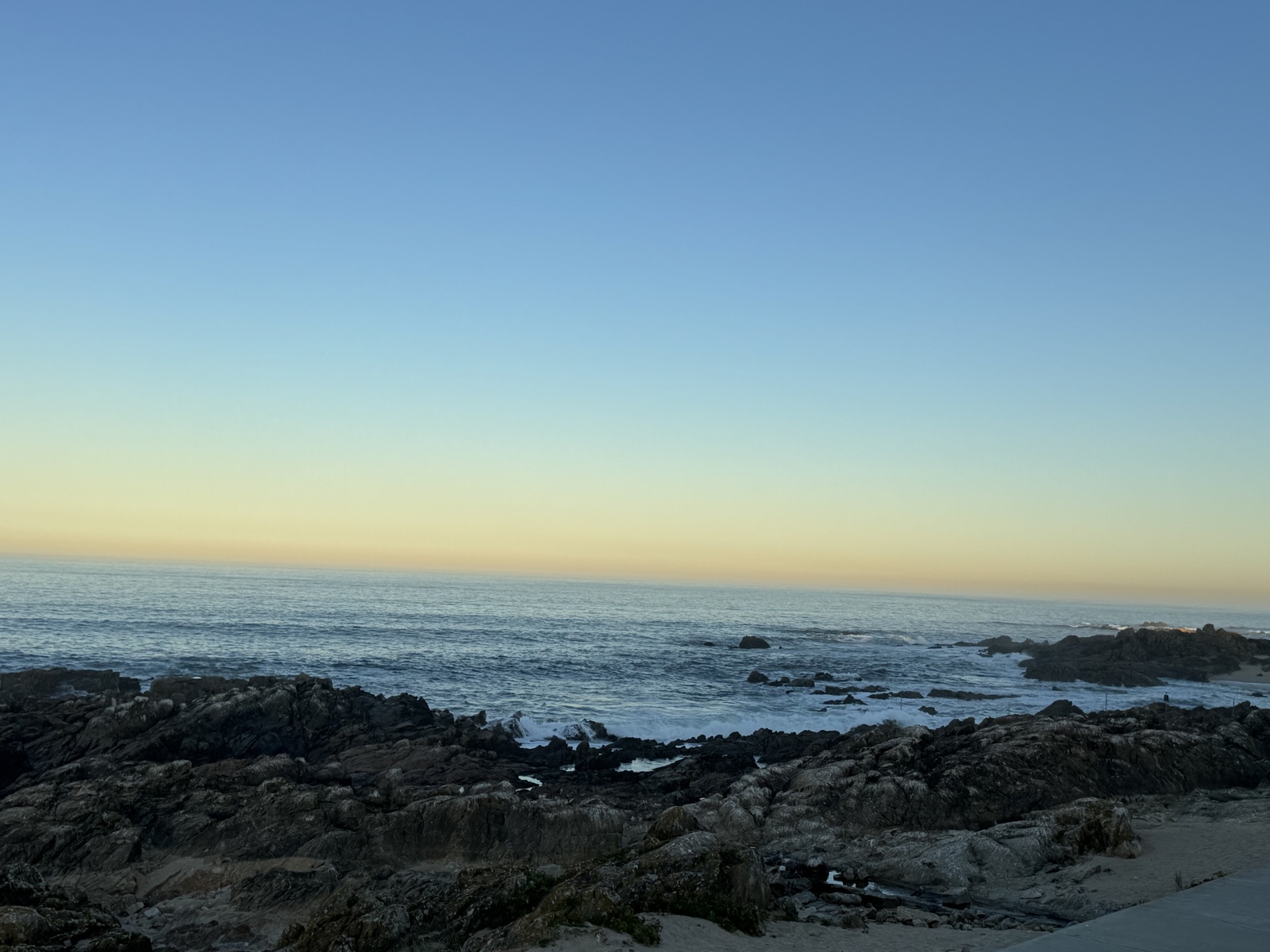 A rocky Atlantic shoreline at first light, pale surf running between dark rocks
          and a wide band of soft yellow on the horizon beneath a clear blue sky.