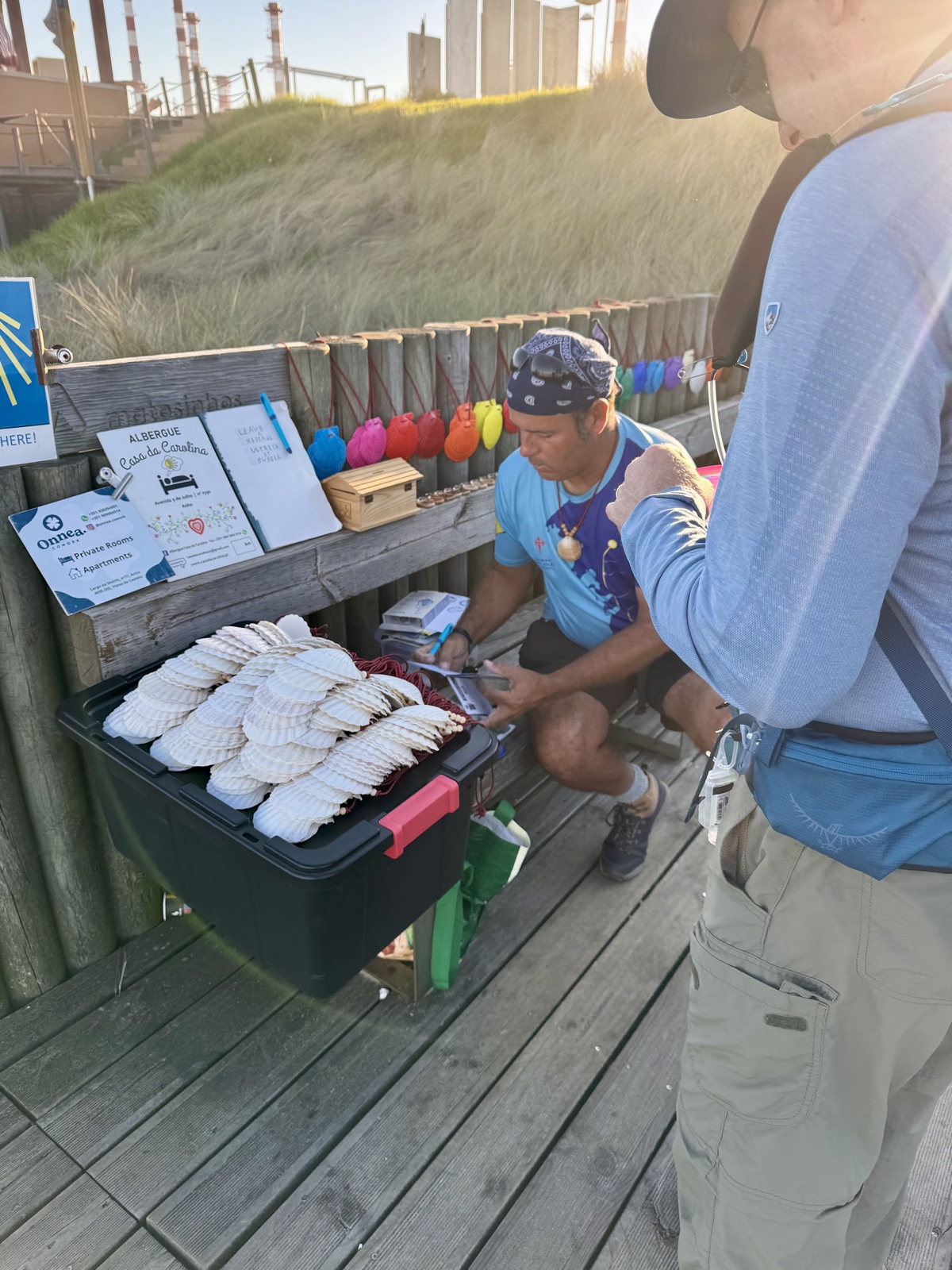 A vendor crouched beside a crate of scallop shells on a seaside boardwalk, a hiker
          standing over it choosing one. Camino signs and coloured buoys hang on the fence behind.