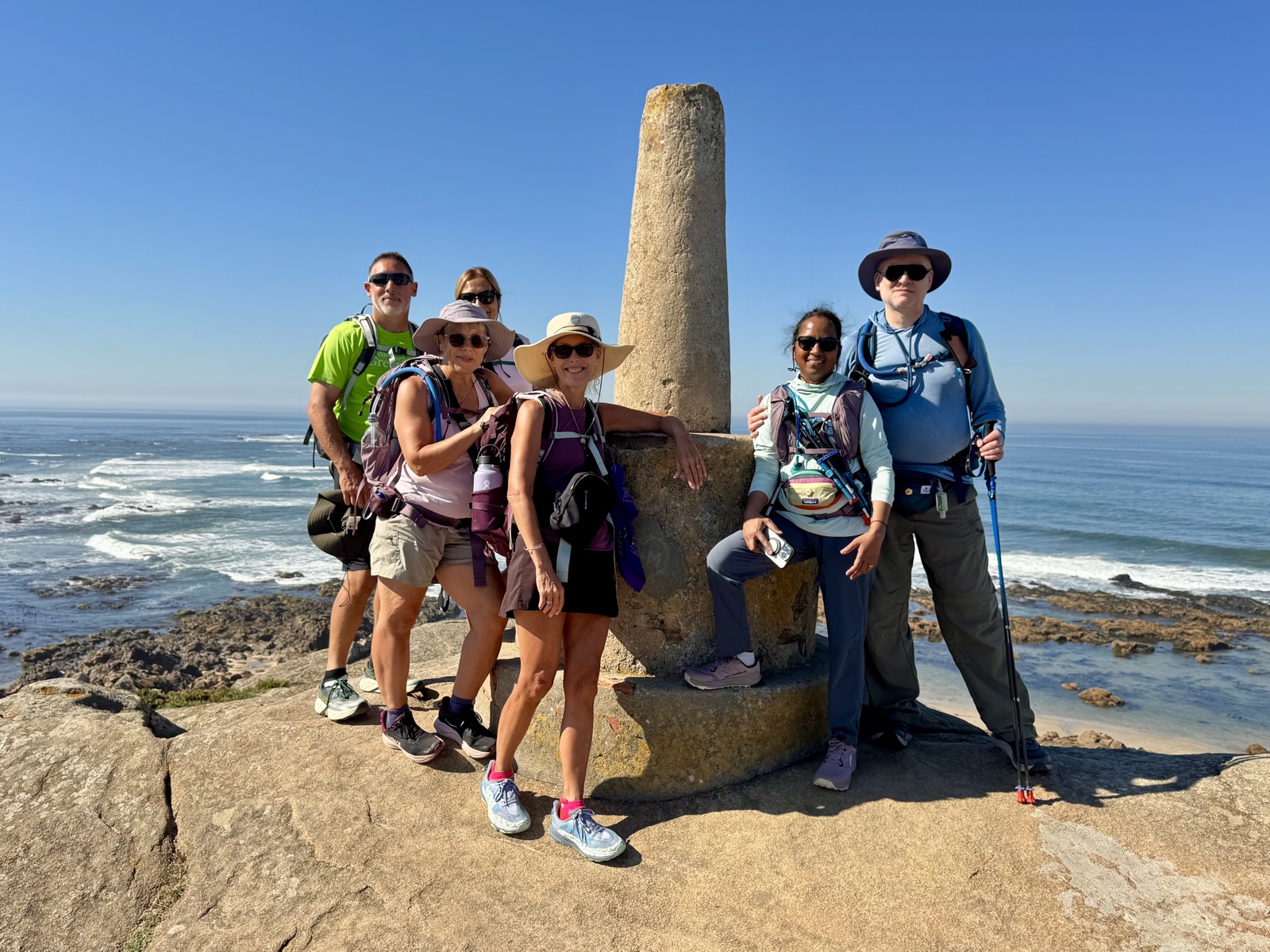 Six hikers in sun hats and daypacks posing around a tall stone obelisk on a
          rocky headland, the Atlantic breaking on the shore behind them under a clear blue sky.