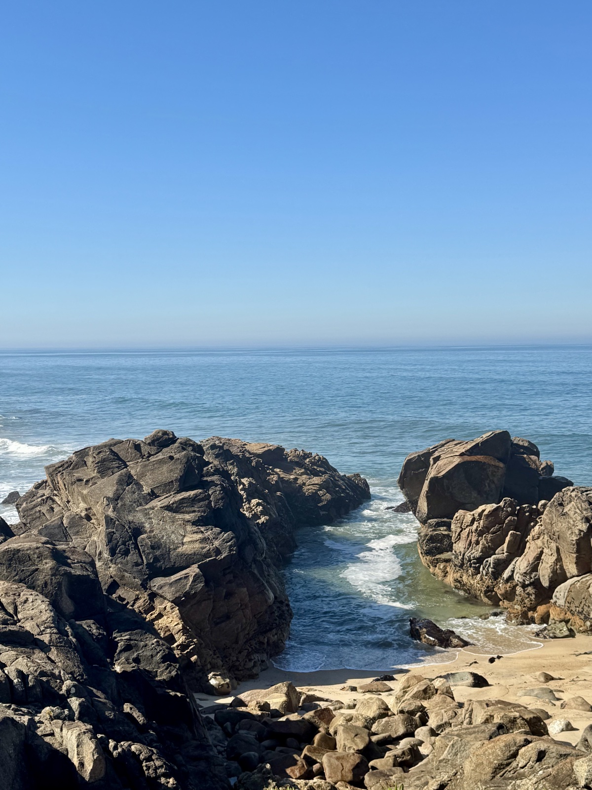 Two large rock outcrops on a sunlit beach with turquoise surf washing into the sandy
          channel between them, the open Atlantic beyond under a cloudless sky.