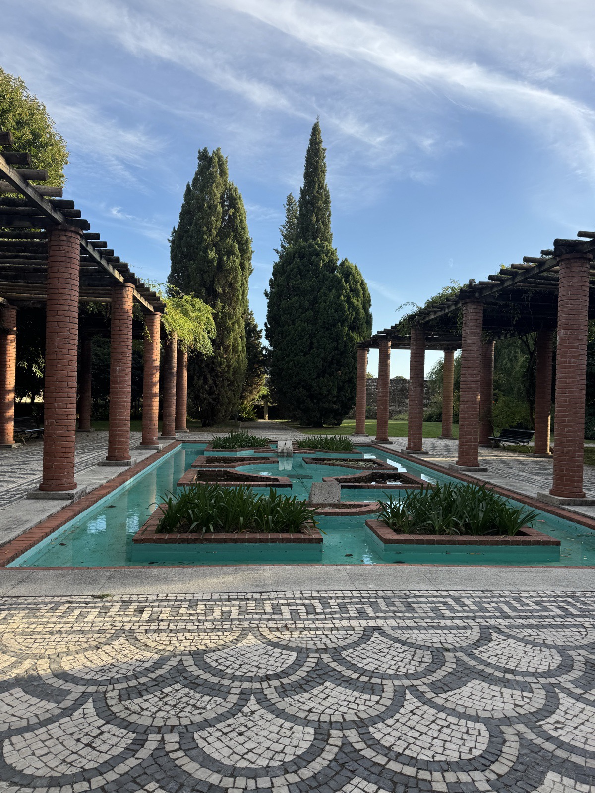 A long rectangular reflecting pool with planted islands, flanked by brick-columned
          pergolas and tall cypress trees, patterned black and white cobblestones in
          front.