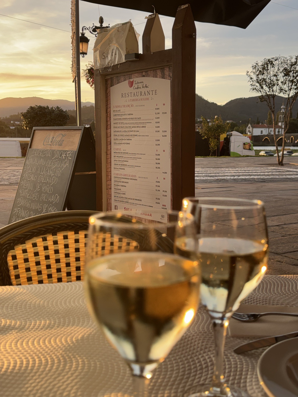 Two glasses of white wine on a café table at dusk, a restaurant menu board and
          chalkboard behind them, hills and a town square beyond.