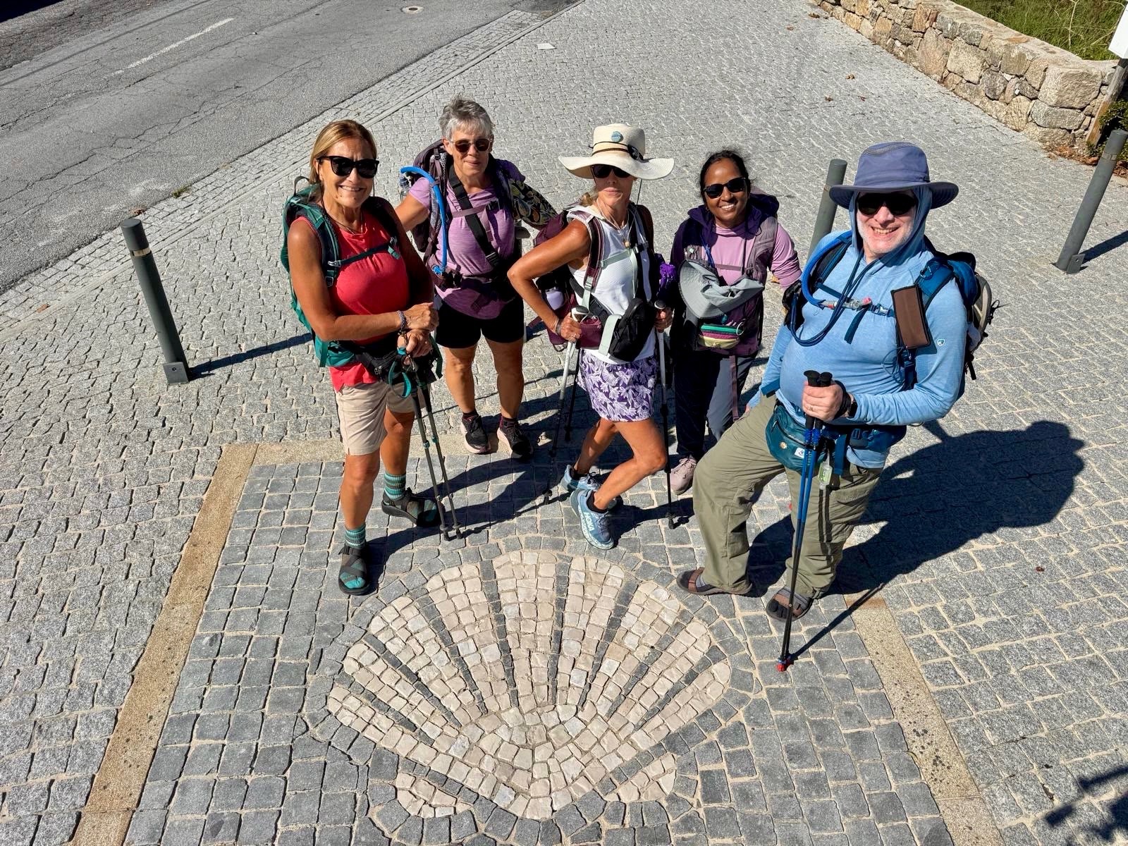 Five hikers with trekking poles and daypacks smiling down at the camera, standing
          around a scallop shell mosaic set into a cobbled pavement.