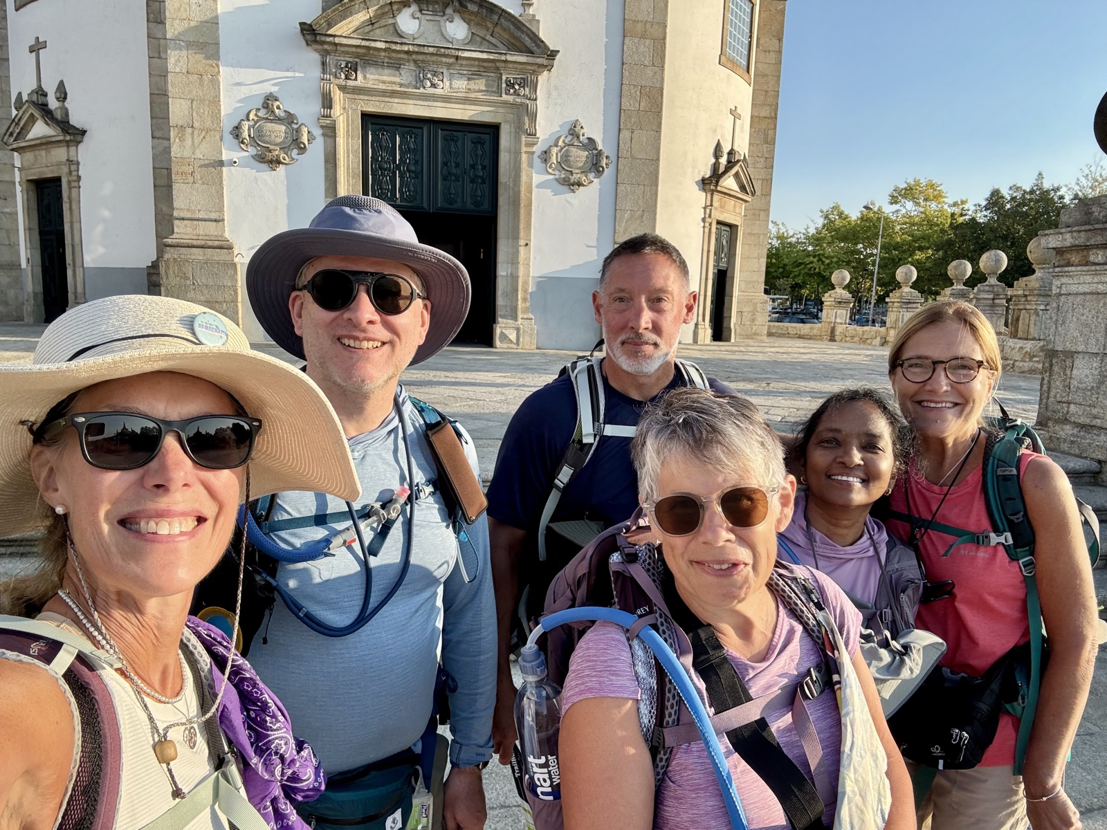 A selfie of six smiling pilgrims in hats and packs in front of an ornate white church in Barcelos.