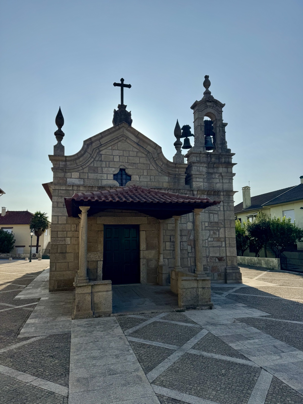 A small granite chapel with a tiled porch roof, a cross and a bell tower, on a paved square under a clear sky.