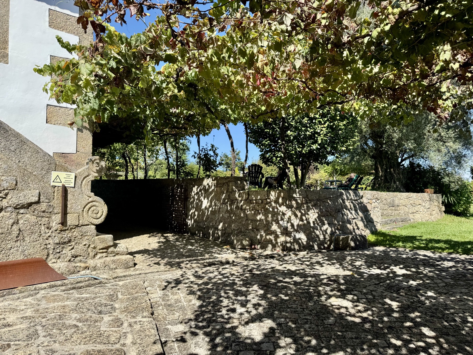 A shaded stone courtyard under a grapevine pergola, a stone wall and green lawn beyond.