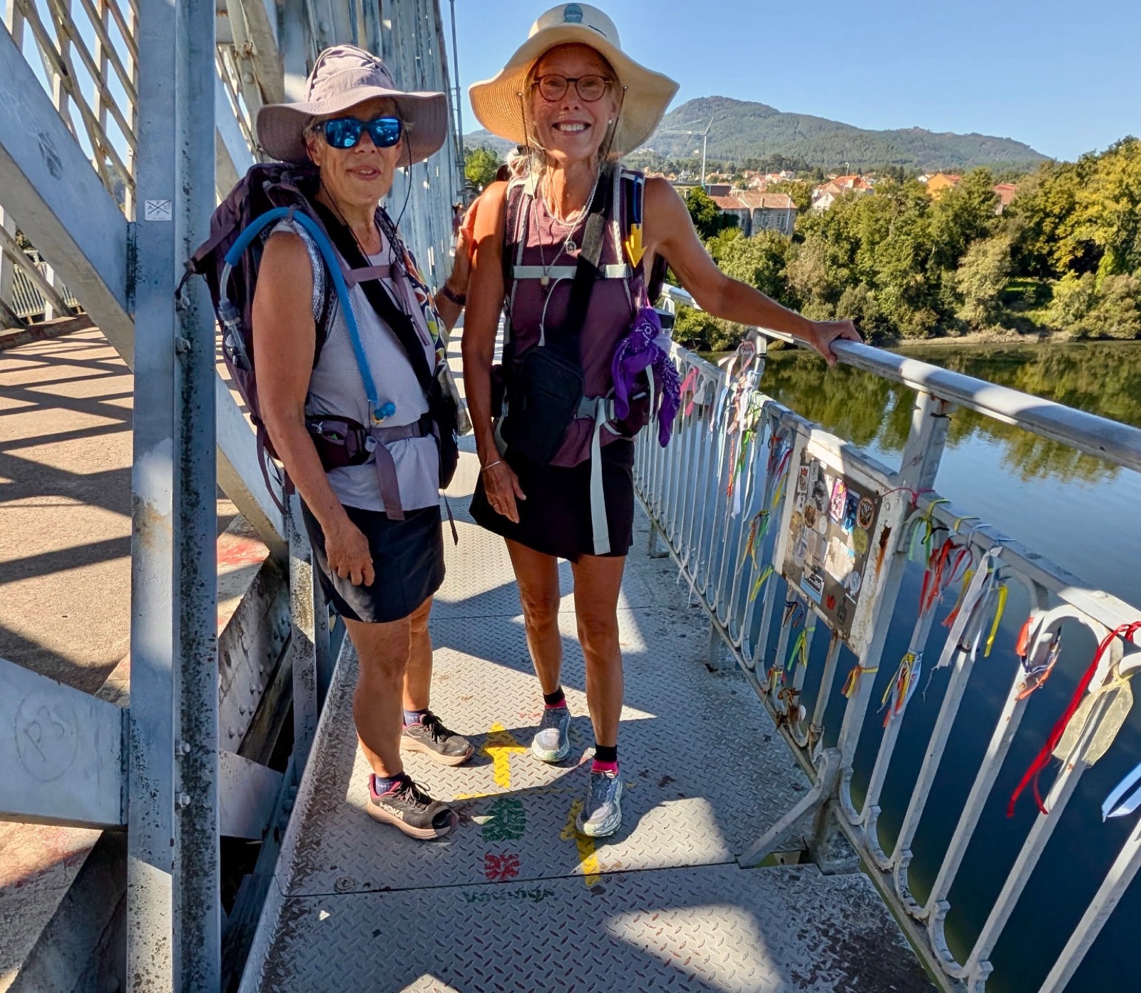 Two pilgrims in sun hats on the walkway of a steel bridge, the railing hung with ribbons, a river and hills behind.