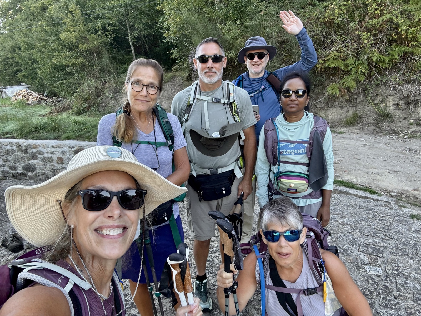Six pilgrims crowd into a morning selfie on a gravel path, one waving.
