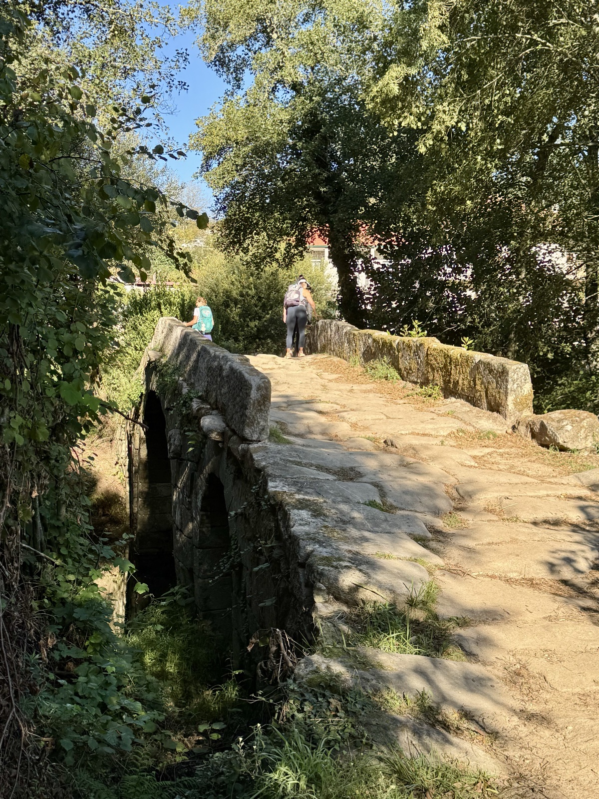 A narrow old stone bridge over a stream, overgrown at the edges, two walkers crossing at the far end.