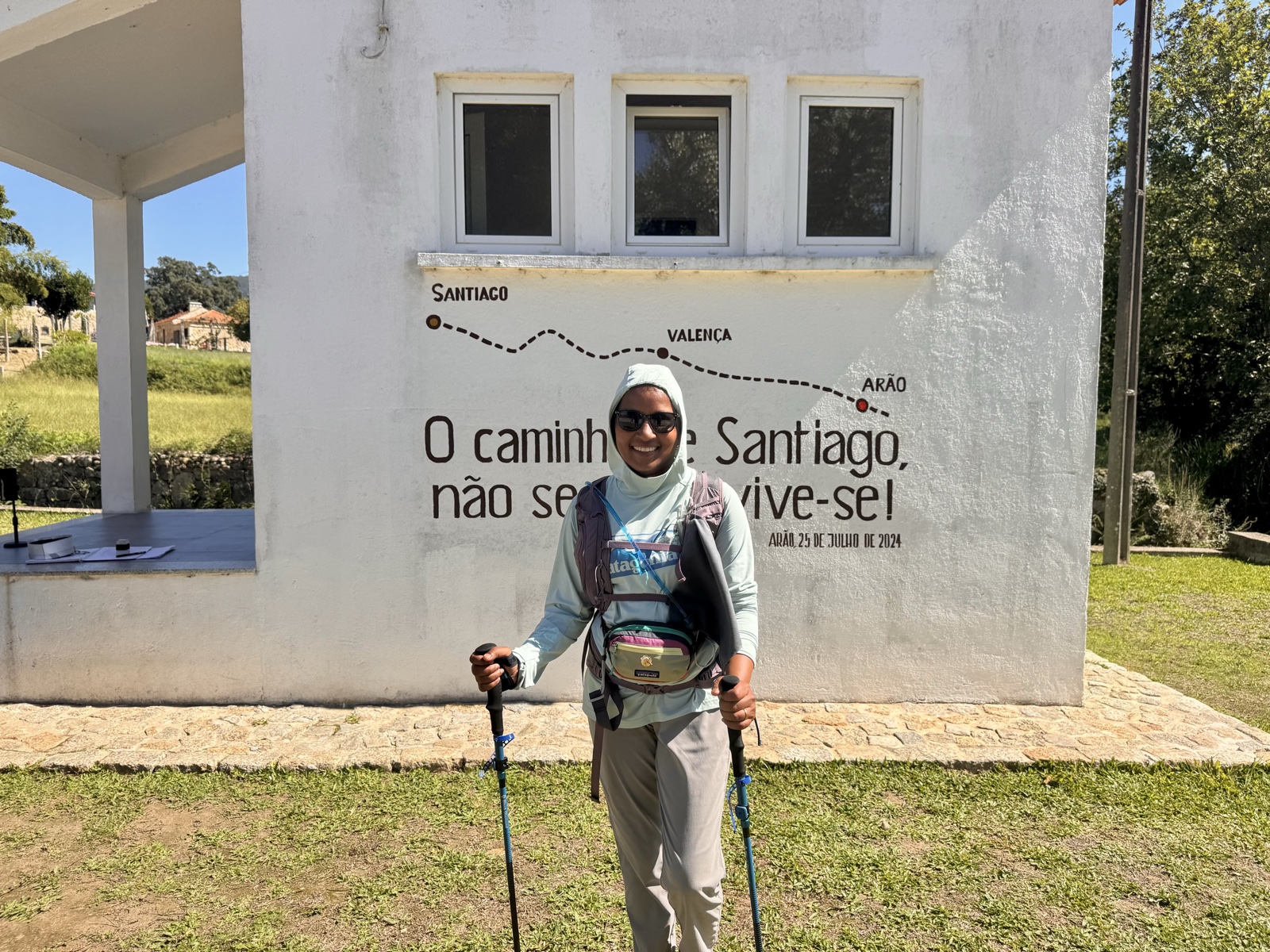 A pilgrim with poles standing before a white wall painted with a Camino route map from Porto to Santiago.