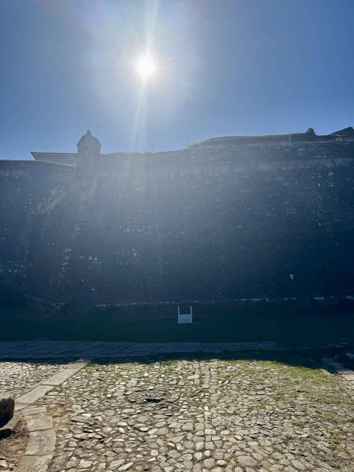 The fortress wall in silhouette against a bright sun, cobbles in the foreground.