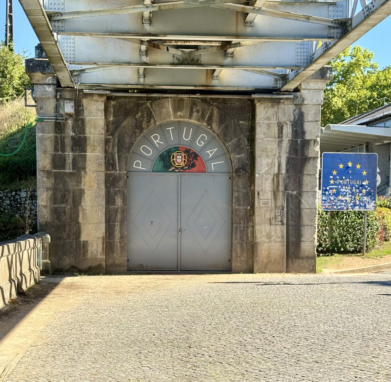 A gate under the steel bridge painted with the word Portugal and its coat of arms, a European Union sign beside it.