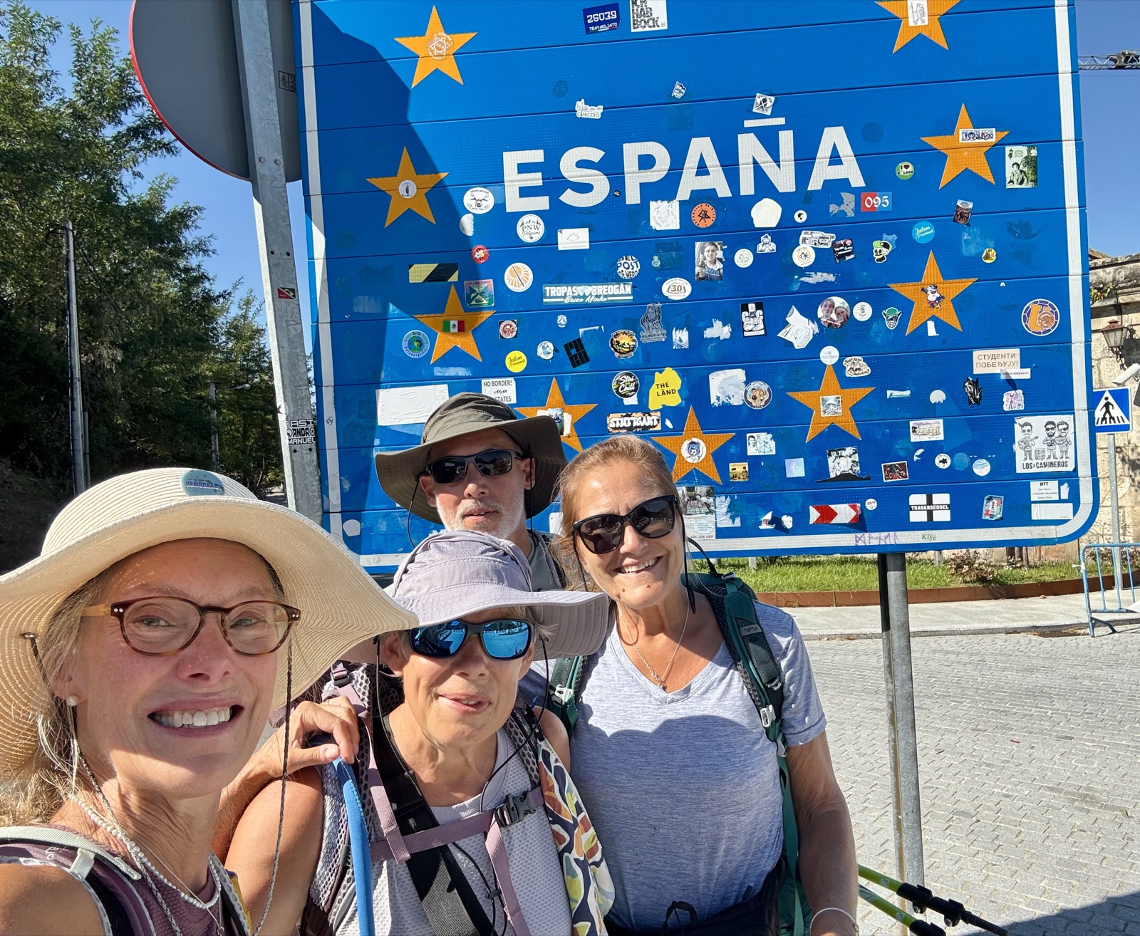 Three pilgrims smiling beneath a blue España border sign covered in stickers.