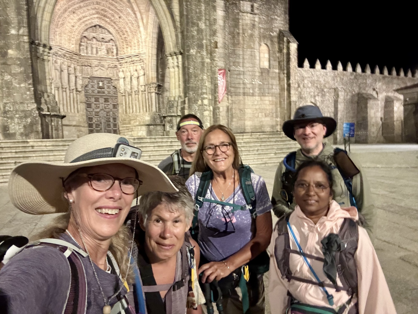 Six pilgrims crowd into a selfie in front of a floodlit medieval cathedral before dawn, packs on and hats pushed back.