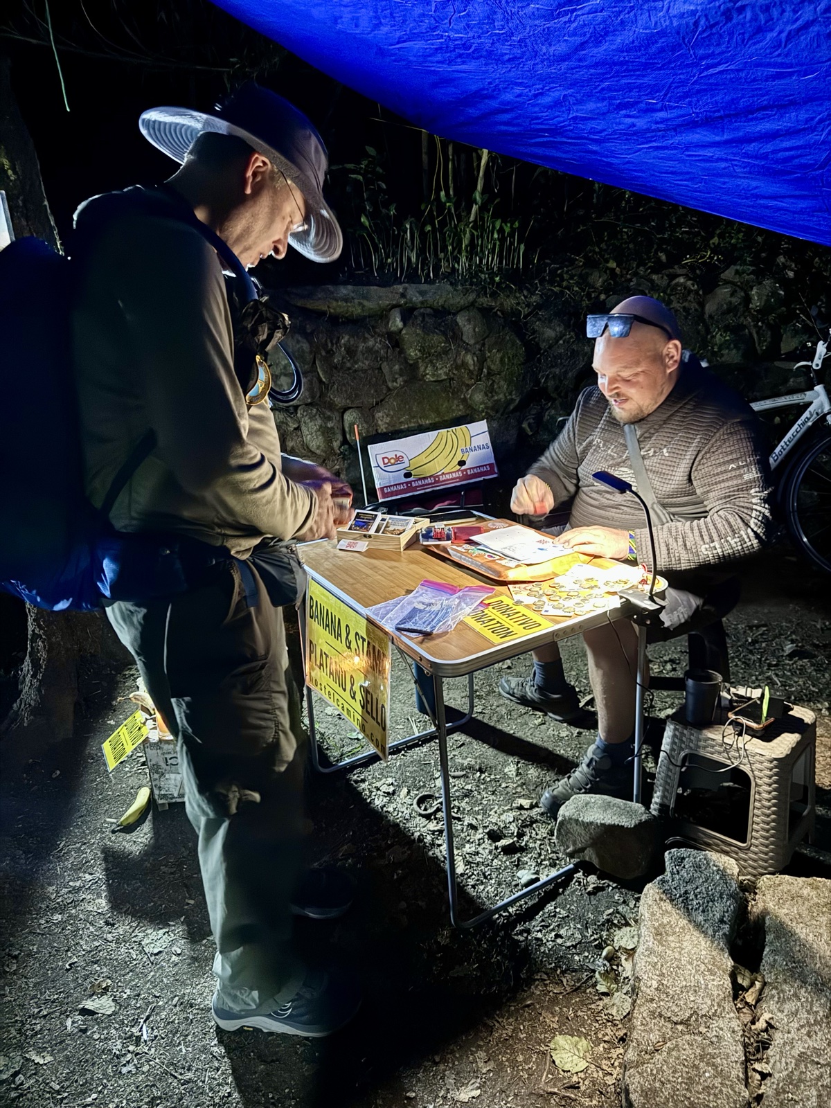 A hiker in the blue half light buying fruit from a man at a small folding stall under a tarp by the trail.