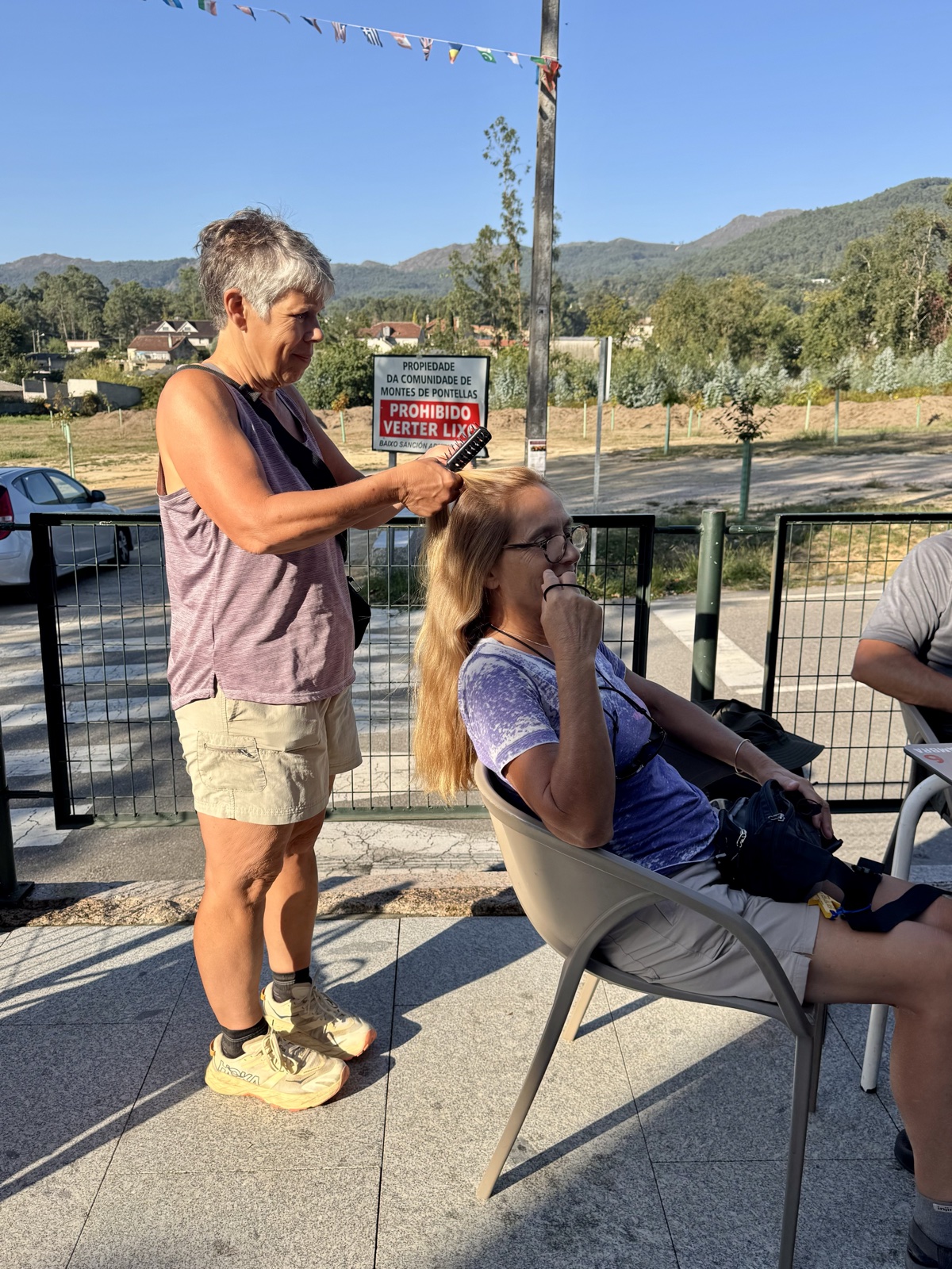 One hiker brushing another's hair as she sits in a plastic chair on a sunny terrace, mountains behind them.