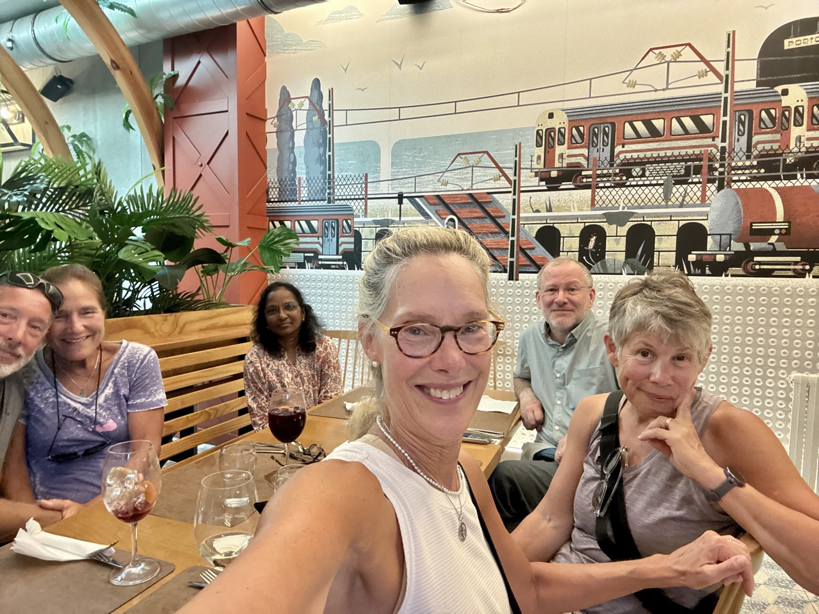 Six friends around a restaurant table raising a selfie, wine glasses in front of them and a painted mural of a train behind.