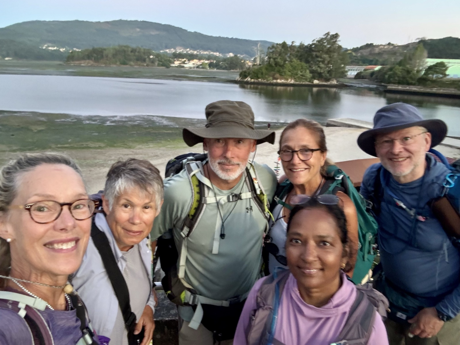 Six pilgrims crowd into a dawn selfie beside a tidal estuary, wooded hills and a village across the water.