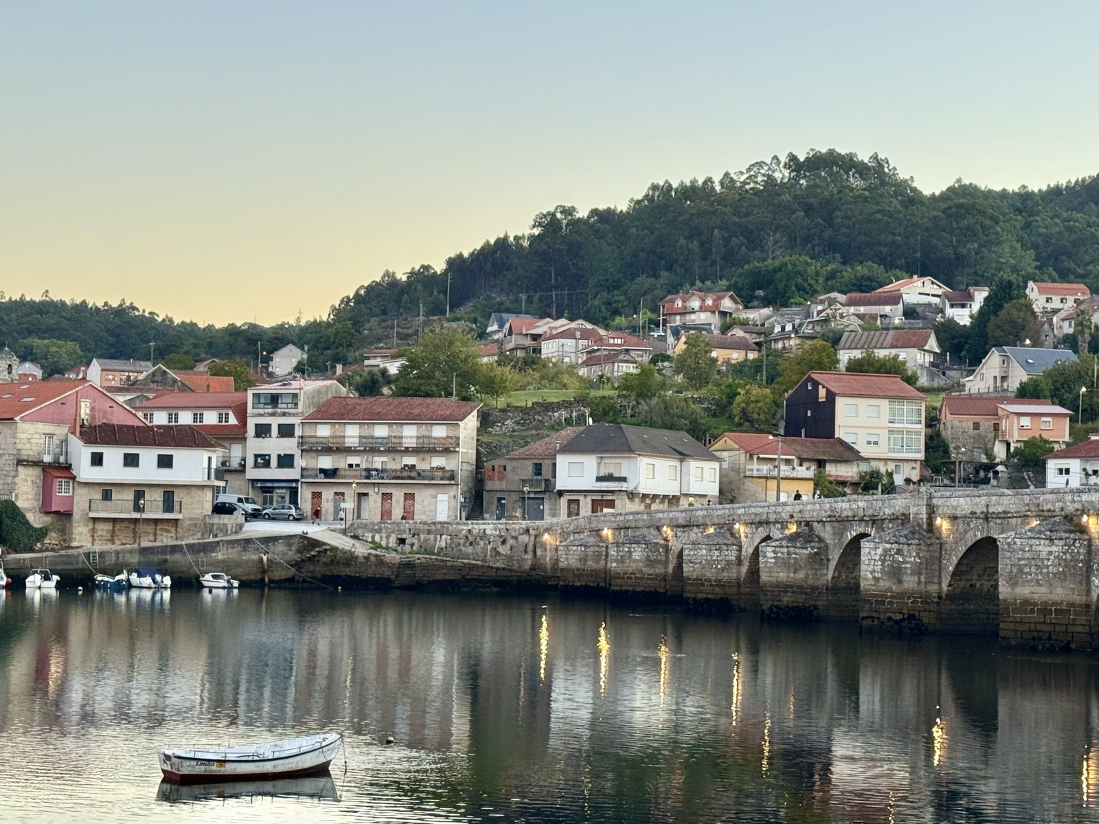 A long medieval stone bridge of many arches crossing still water at dawn, a small rowboat in the foreground and the village of Arcade on the hill behind.