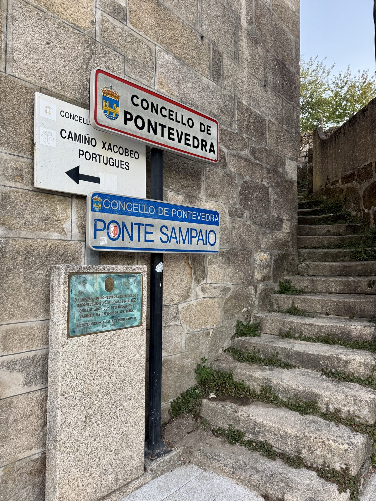 Road signs on a granite wall reading Concello de Pontevedra and Ponte Sampaio, an arrow for the Portuguese Camino, beside a flight of worn stone steps.