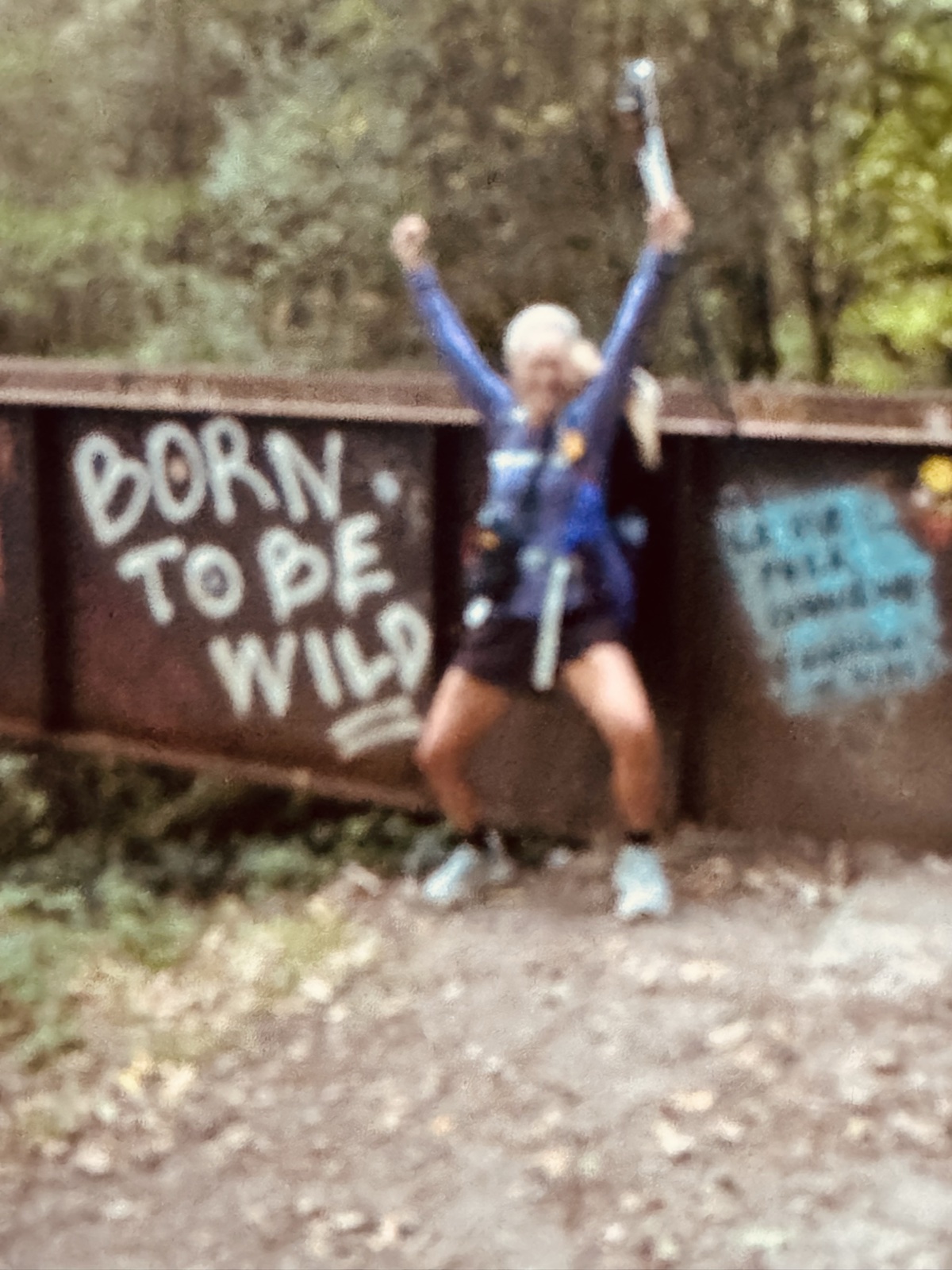 A pilgrim with arms and a trekking pole raised in triumph in front of a rusted steel barrier painted Born to be Wild.
