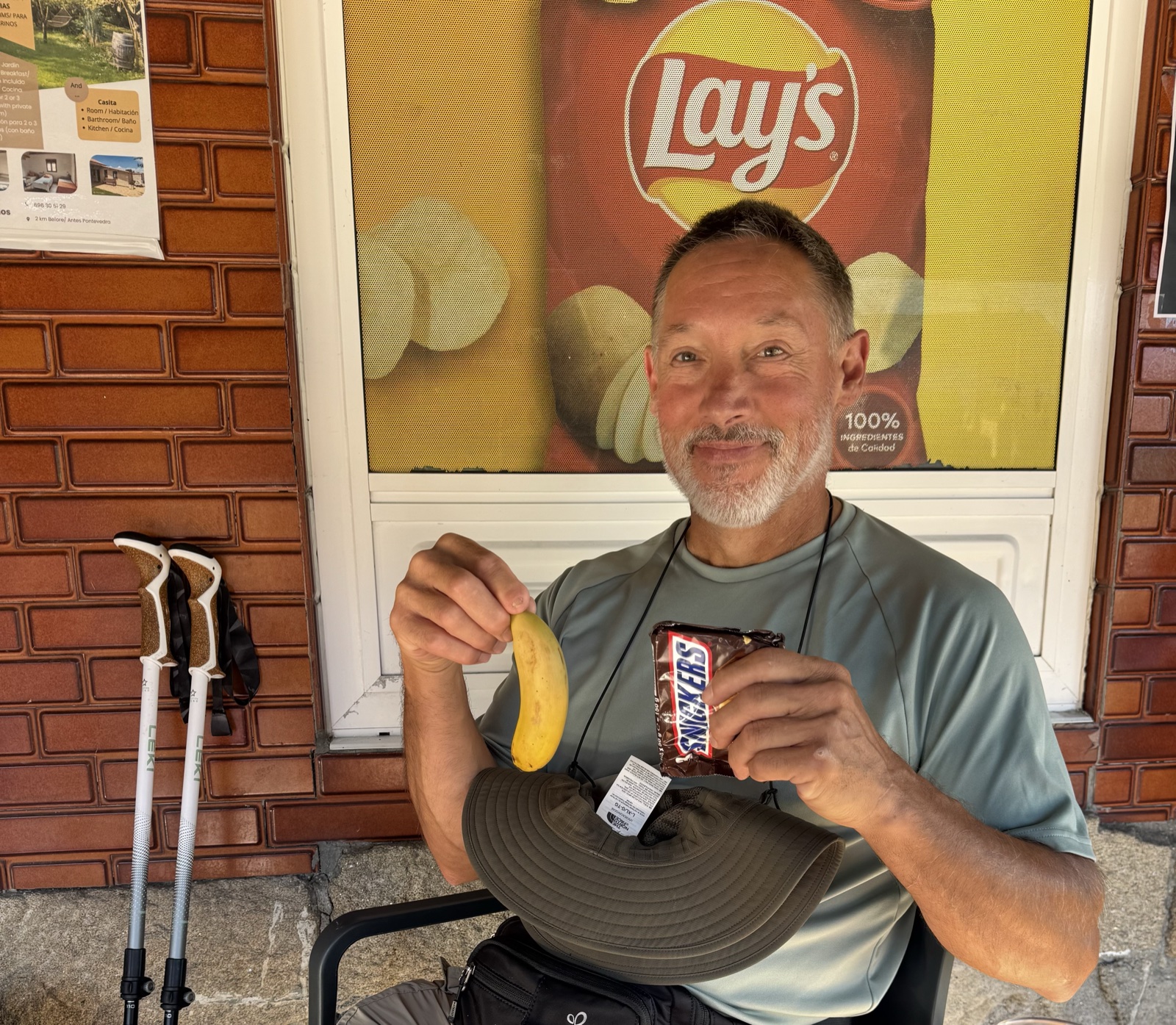 A smiling hiker holding a banana in one hand and a Snickers bar in the other, seated outside a cafe under a Lay's crisps poster.