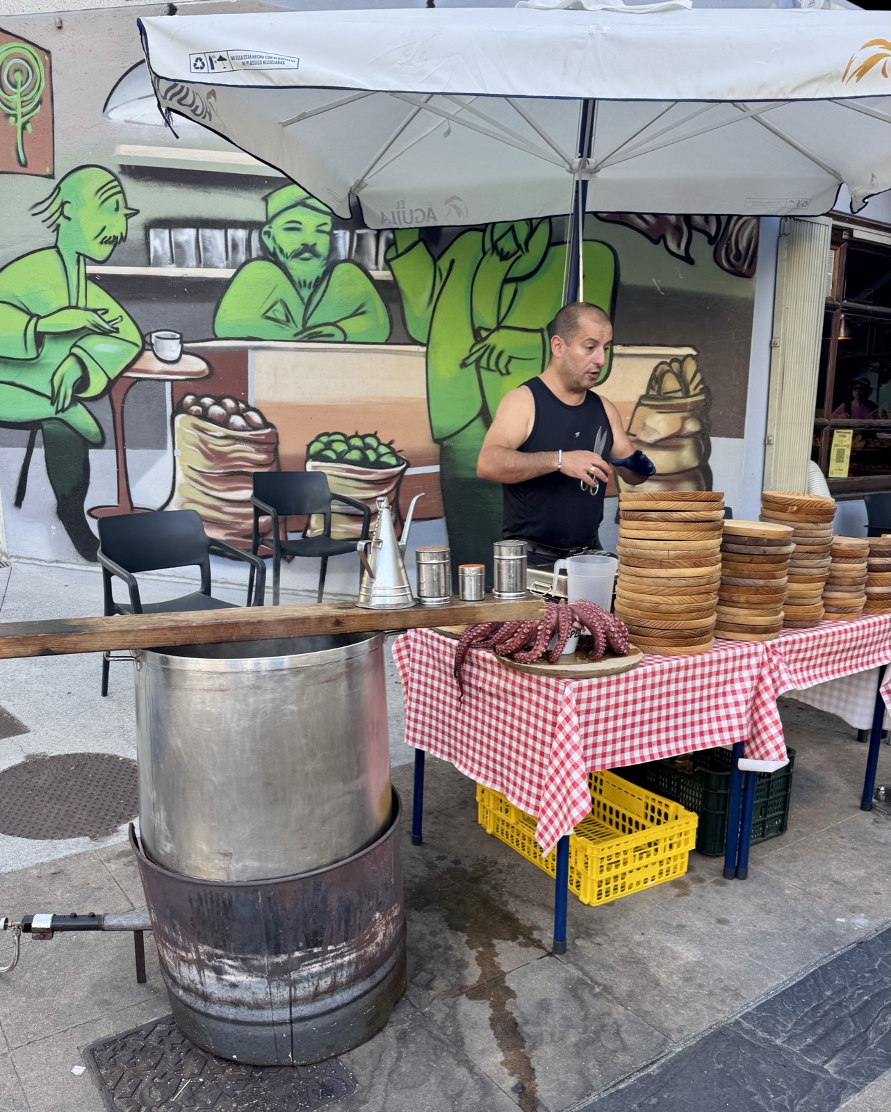 A man at a street stall with red checked tablecloths and stacks of wooden plates, a large steel octopus cooking pot in front, a mural of green figures behind.