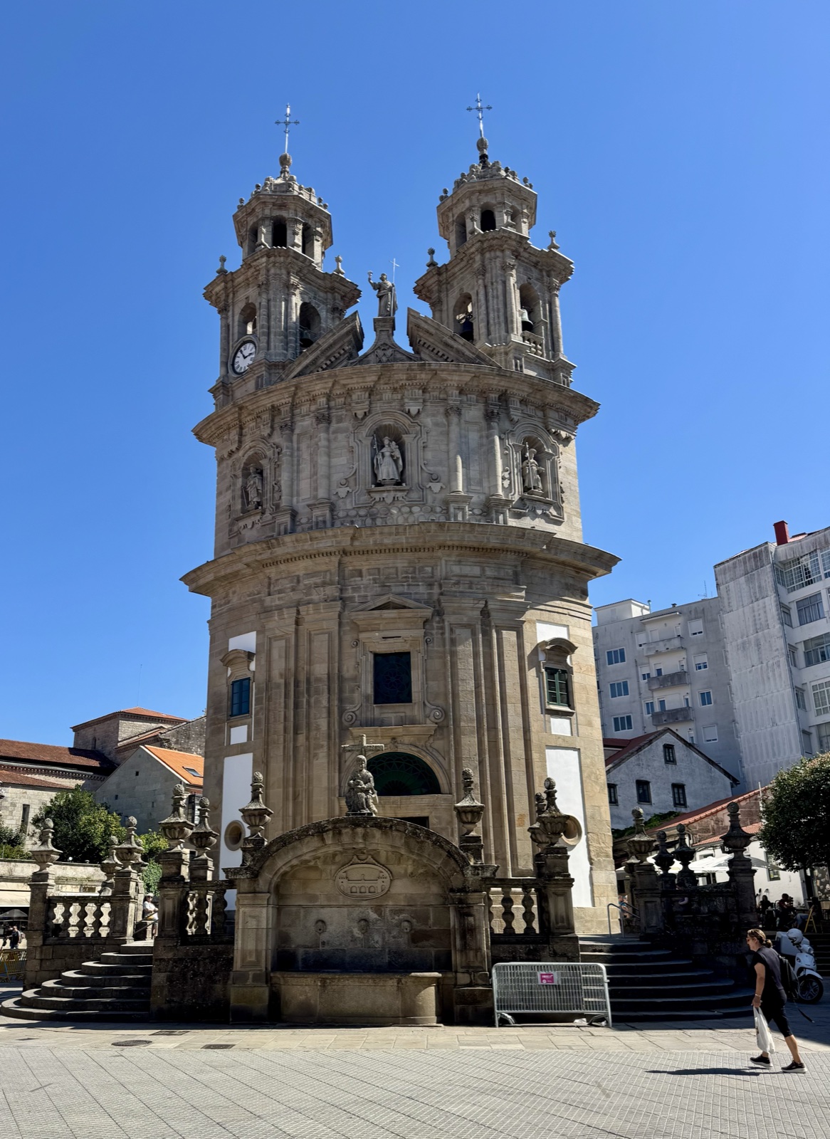 The Pilgrim Virgin church in Pontevedra, a tall curved baroque front with twin bell towers and a fountain, under a clear blue sky.