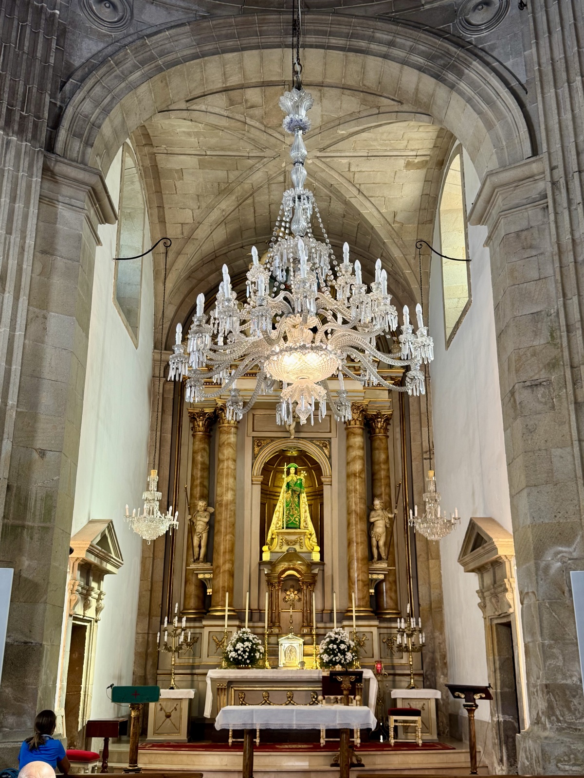 The nave of a stone church, a large crystal chandelier hanging before a gilded altar with a statue of the Virgin.