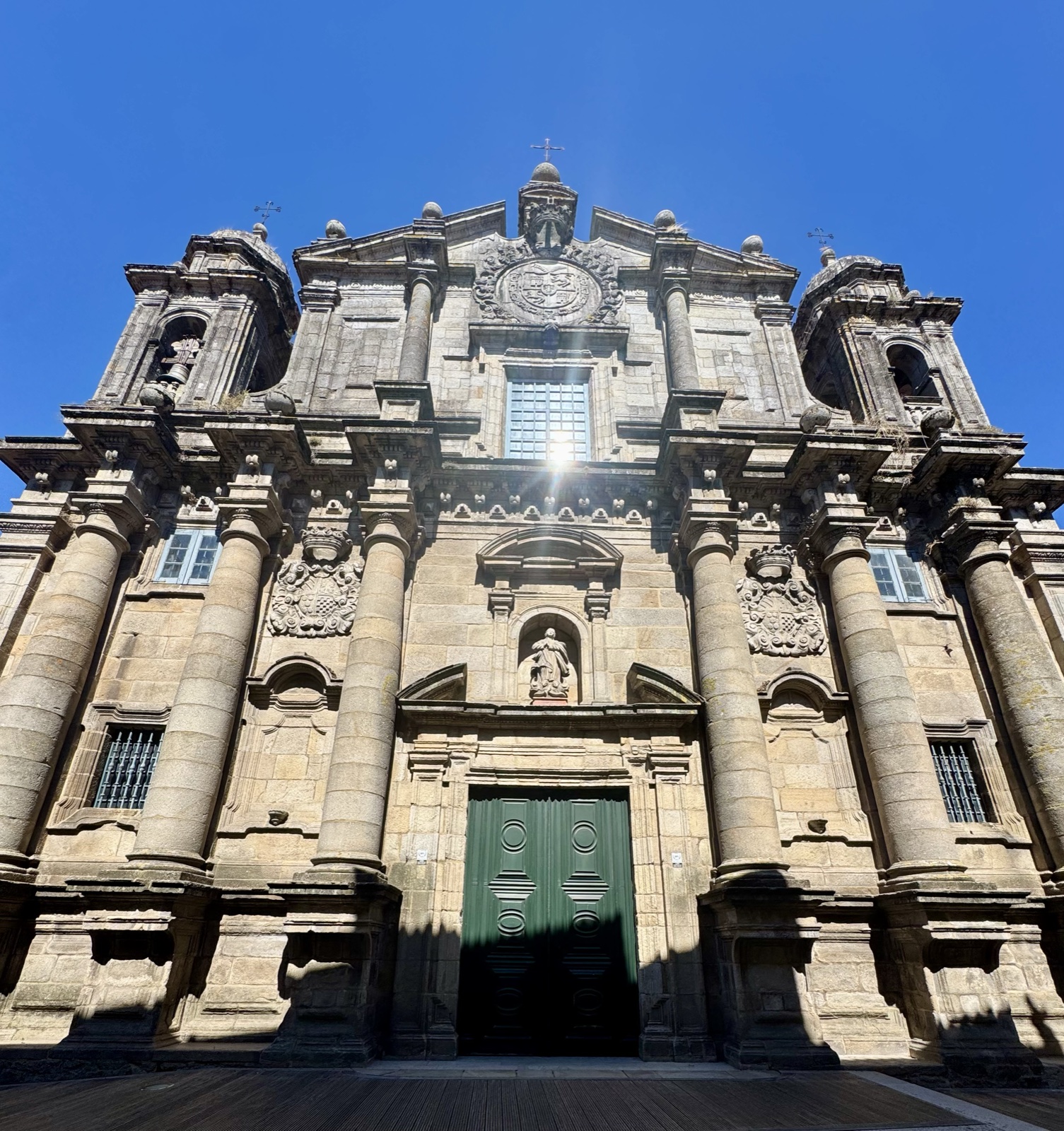 A grand baroque granite church front seen from below, tall columns, carved crests and a green double door, the sun flaring behind a window.