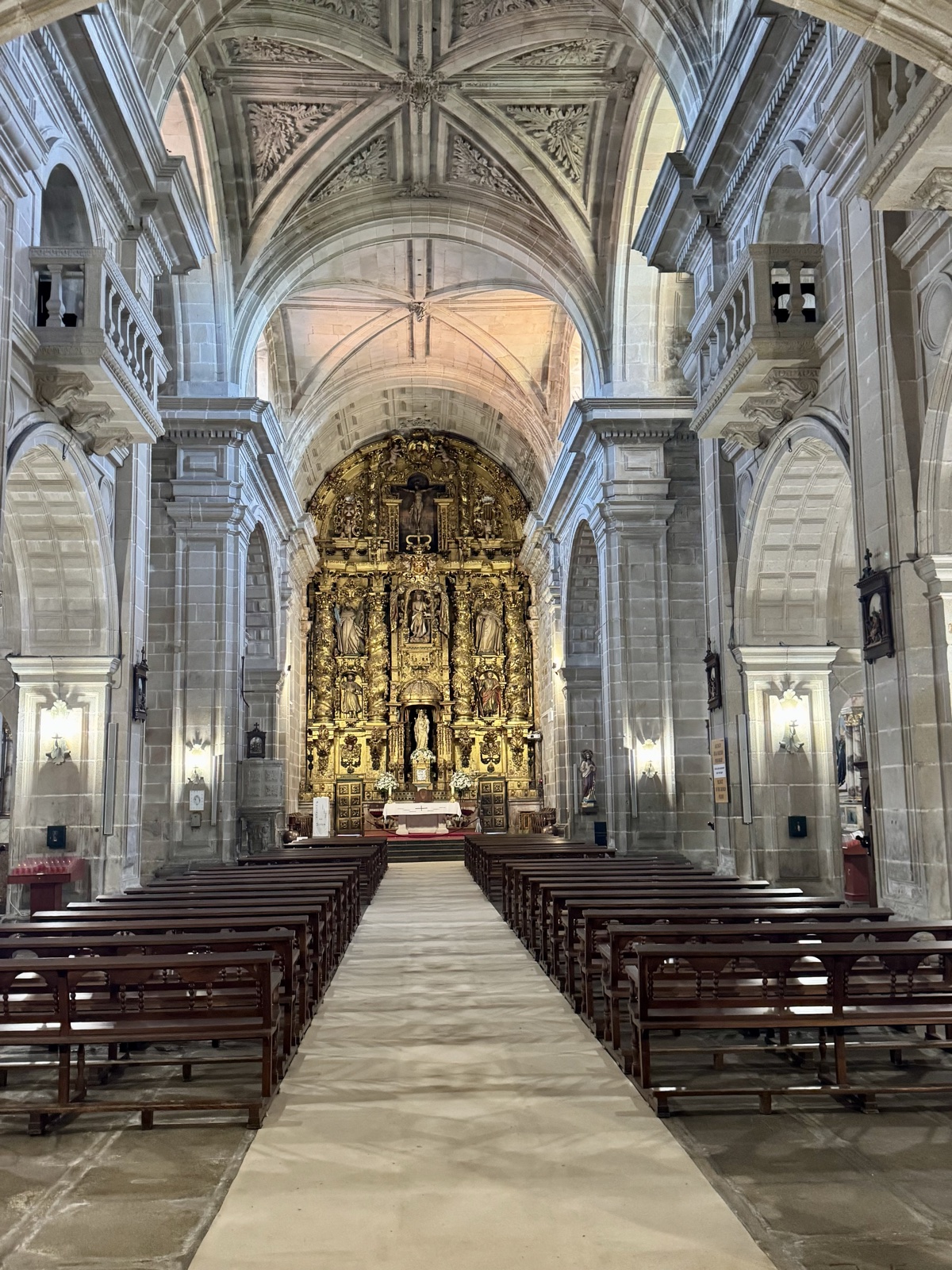 The long nave of the monastery church, rows of wooden pews leading to a gilded altarpiece under a carved stone vault.