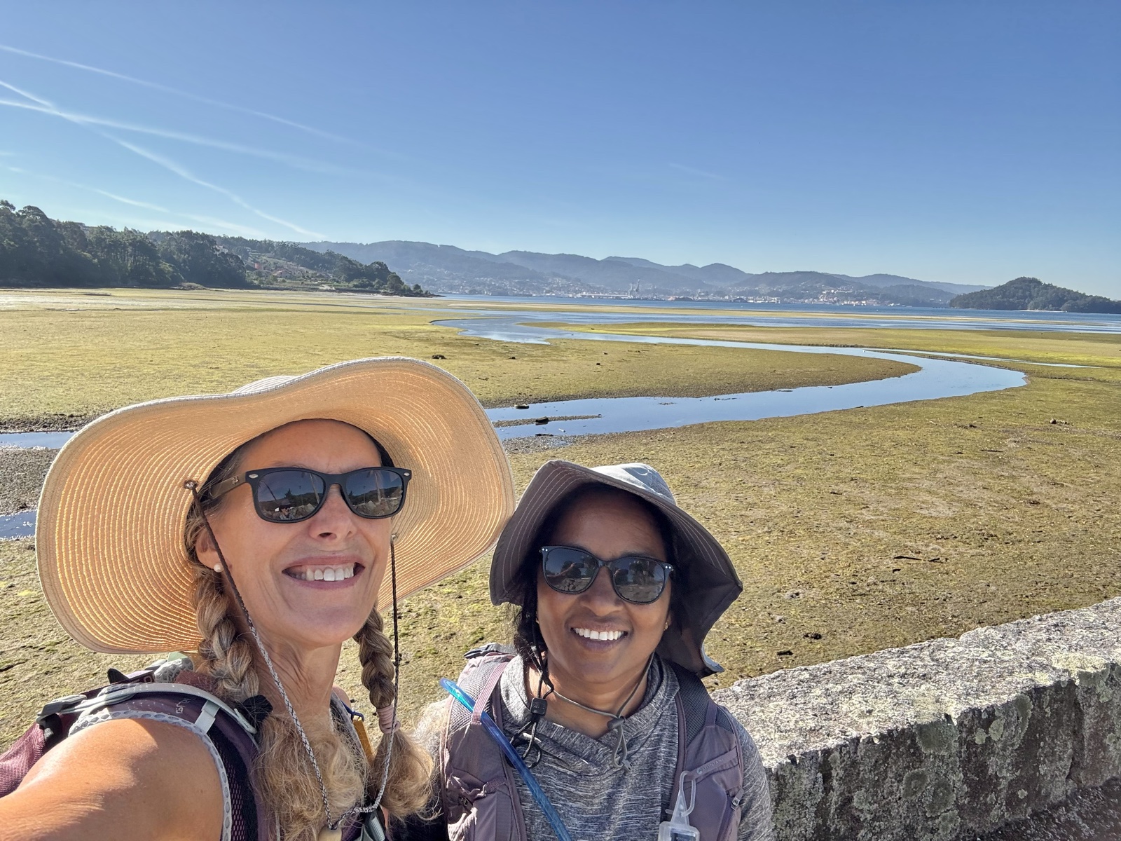 Two pilgrims in sun hats smiling in a selfie above wide tidal flats, mountains across the water.