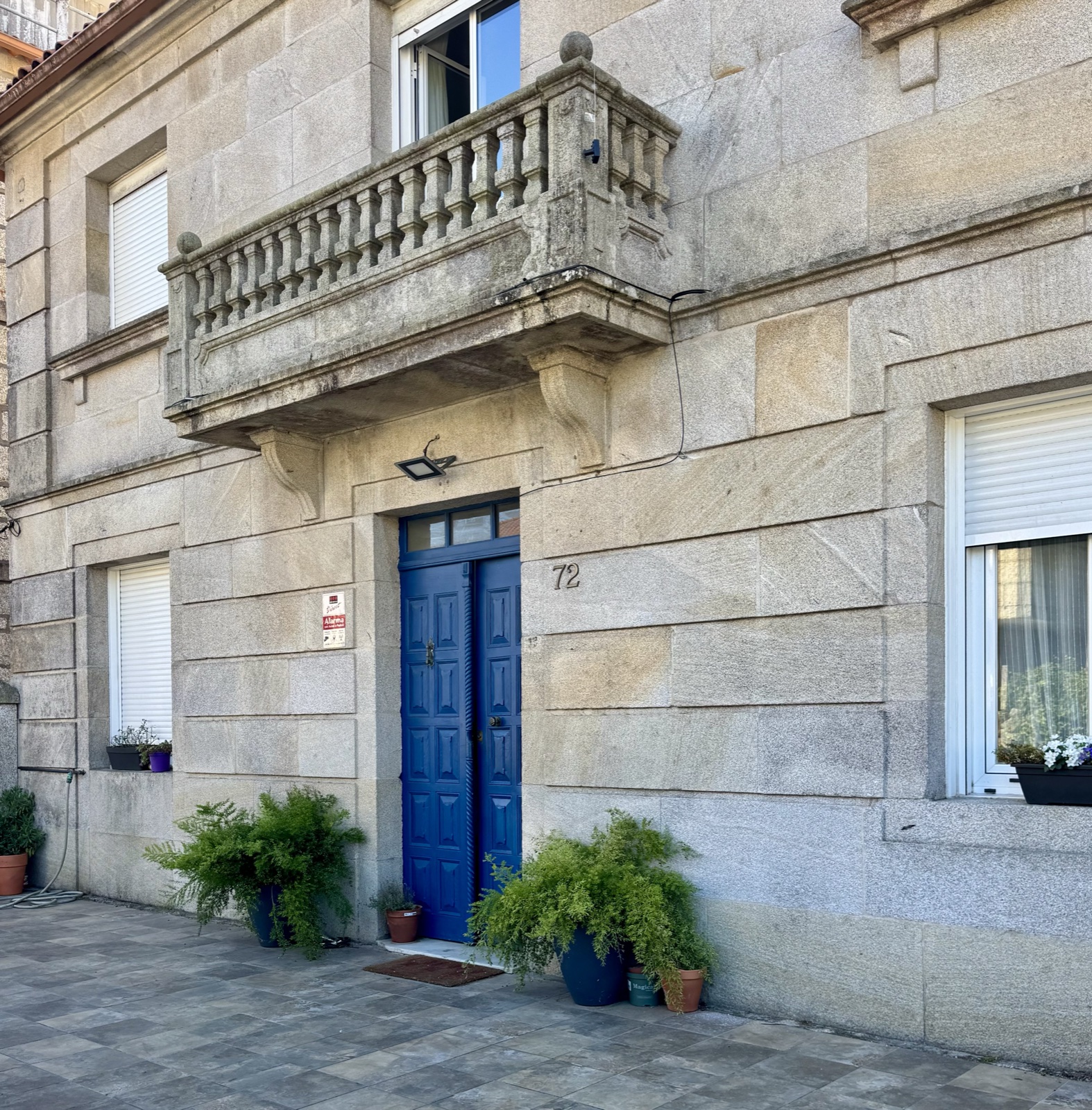 A granite house with a blue door, a stone balcony above it and ferns in pots at the step.