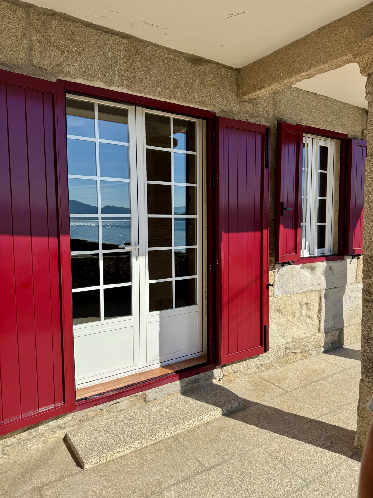 White French doors framed by red shutters in a granite wall, the sea reflected in the glass.