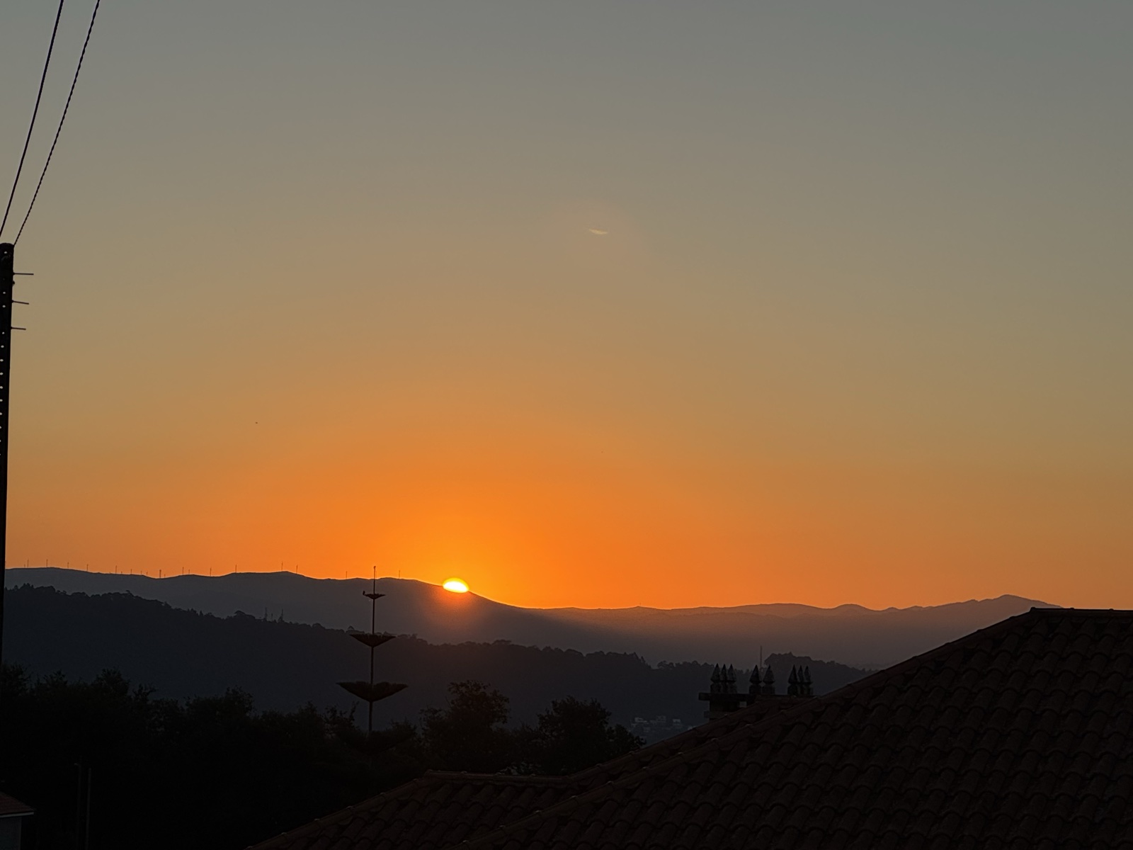 The sun just breaking over a dark mountain ridge in an orange sky, rooftops and a utility pole in silhouette.