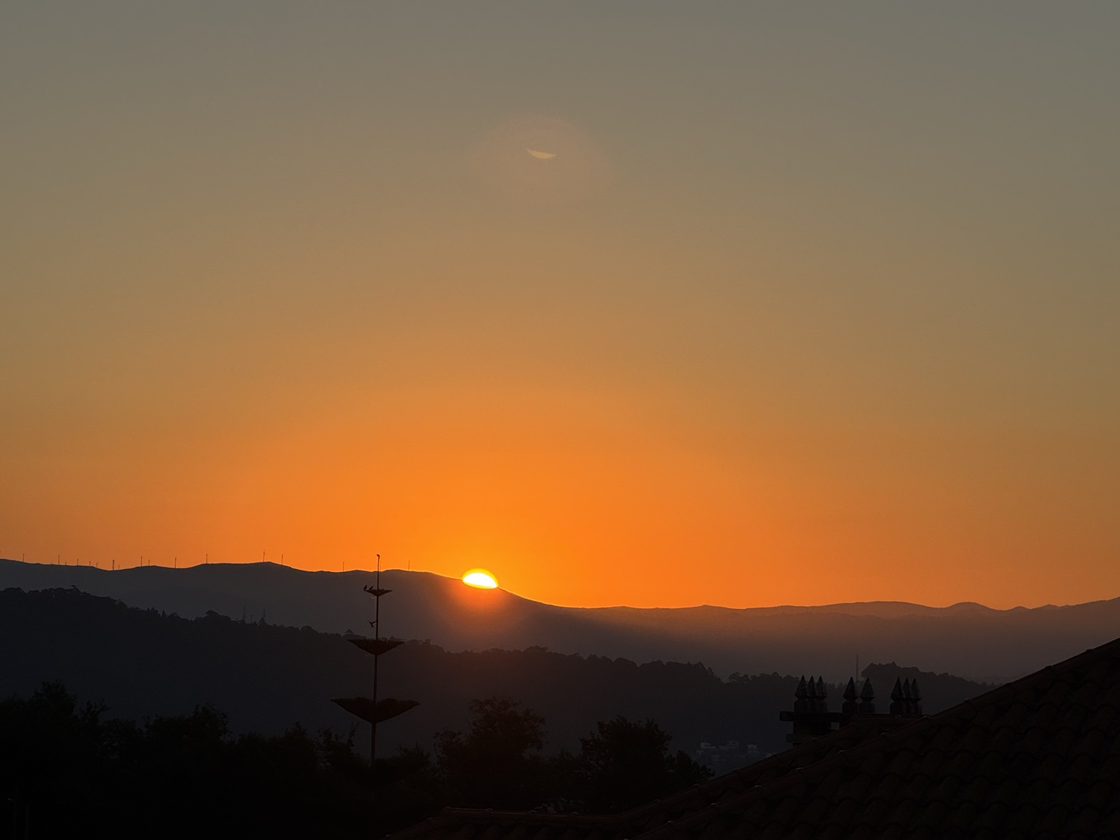 The sun rising over the mountains in an orange sky, with rooftops in silhouette below.