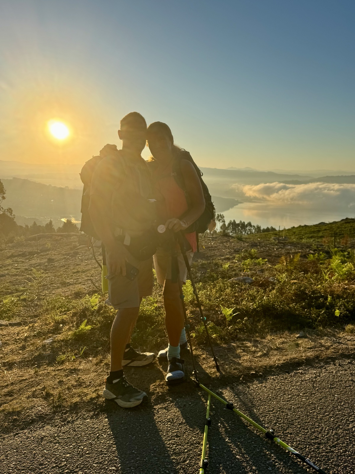 Two hikers with packs and poles standing close together, backlit by the rising sun above a valley of cloud.