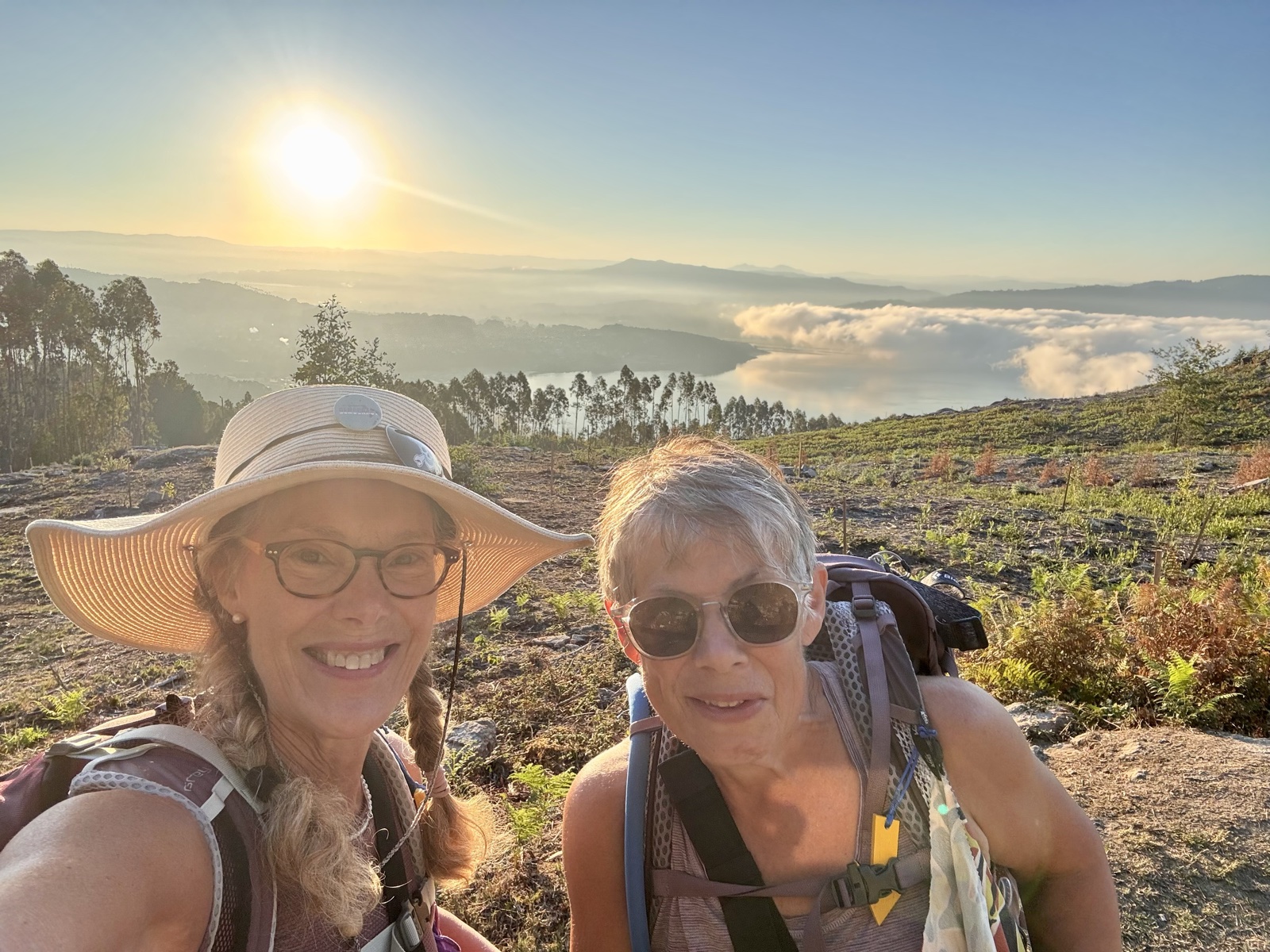 A selfie of two smiling women in hats and sunglasses on a hillside, the sun and a cloud-filled valley behind them.