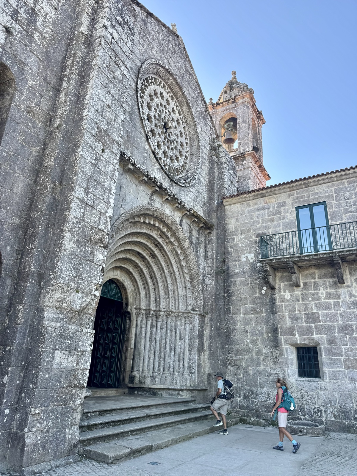 The stone facade of the monastery church, with a round rose window above an arched doorway and two pilgrims walking past.