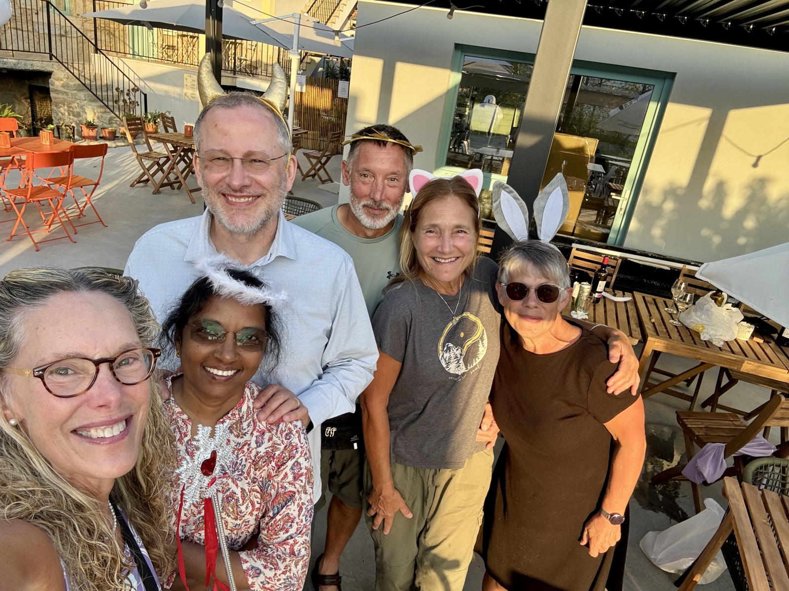 A group selfie of six smiling friends on a sunny terrace, several wearing party animal ears and horns.