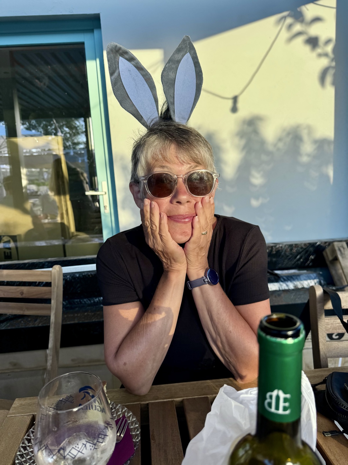 A woman in bunny ears and sunglasses resting her chin in her hands at an outdoor table, a wine bottle in front of her.