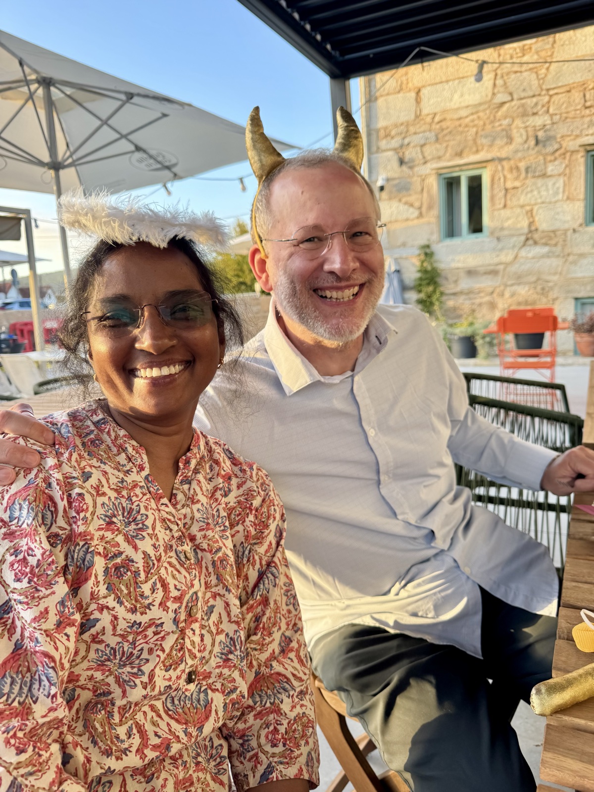 A smiling woman in a floral top and a man wearing golden horns, seated together on a terrace.
