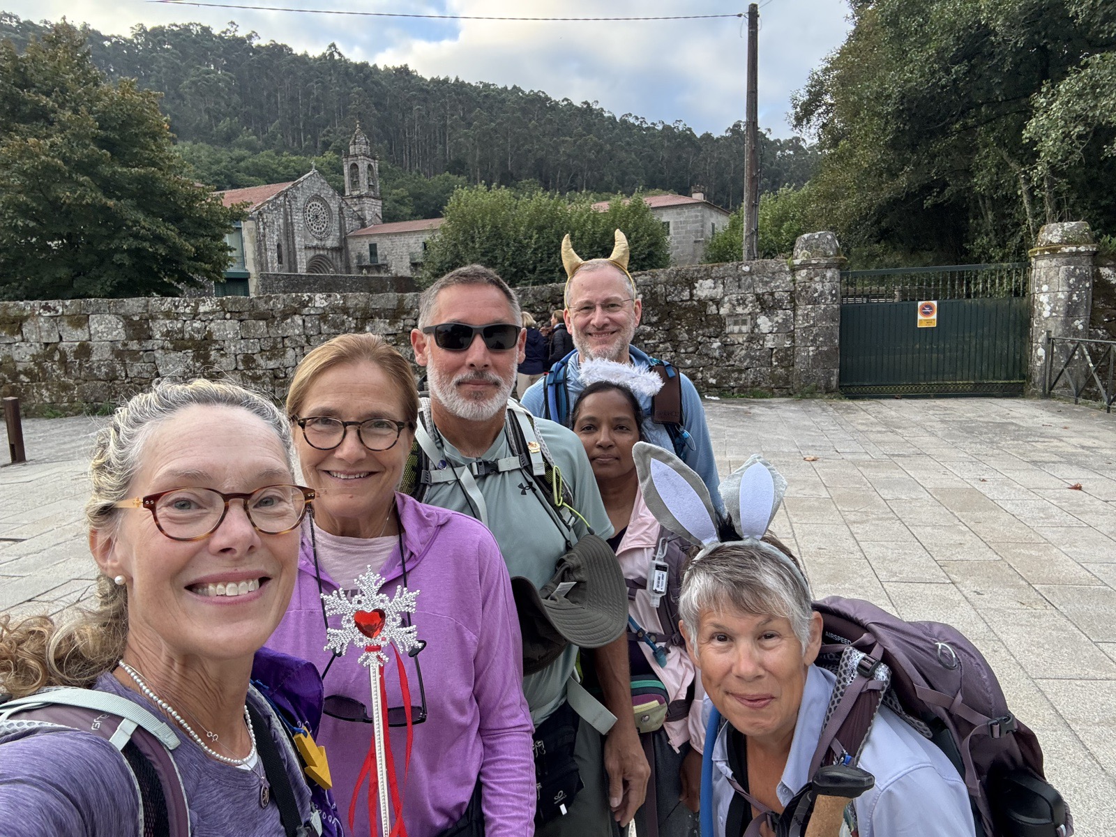 A selfie of six smiling pilgrims with packs in a stone-paved square, a granite church on the hillside behind them.