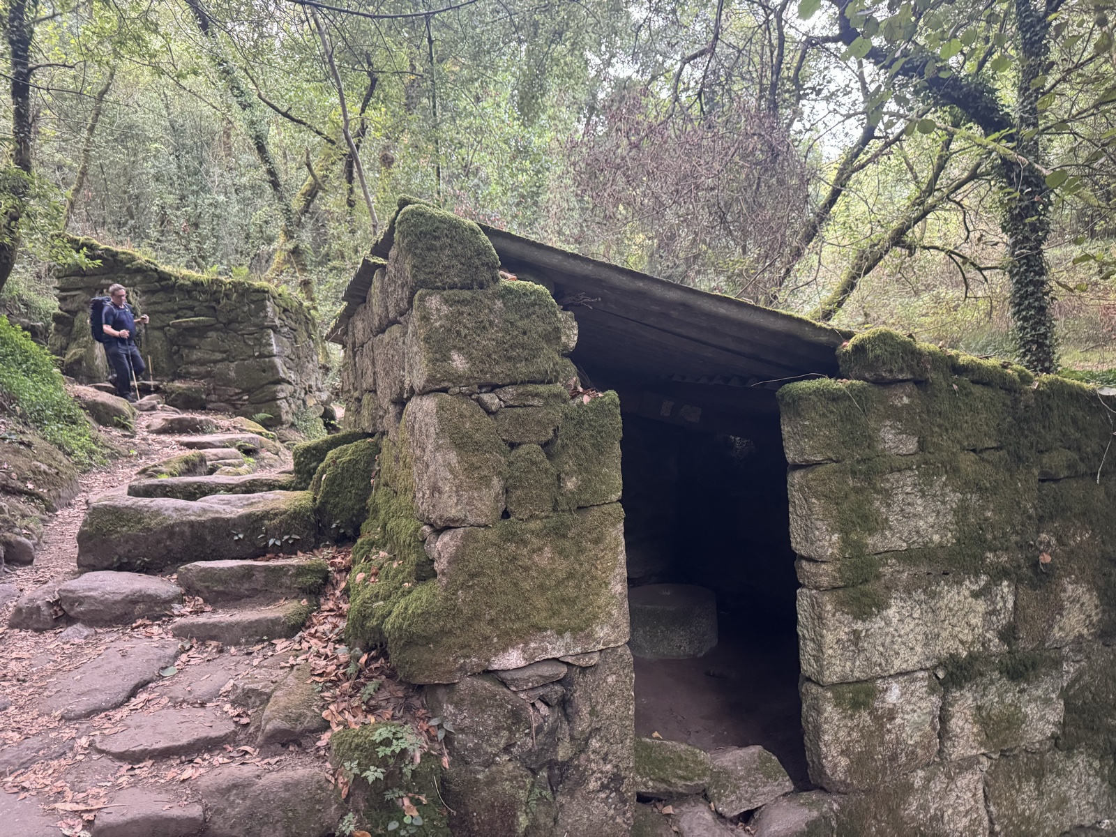 A roofed stone mill building beside mossy stone steps, a hiker climbing the path behind it.