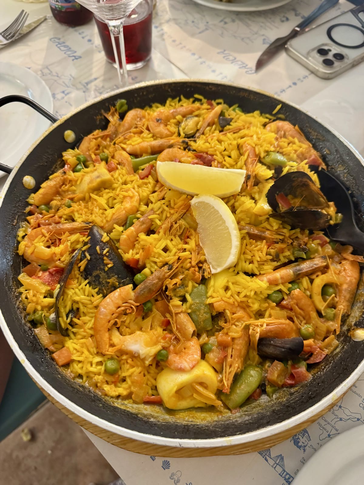 A pan of seafood paella with prawns, mussels, peas, and two lemon wedges on a restaurant table.