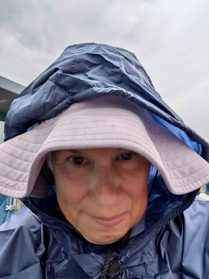 A pilgrim in a navy rain hood and a lavender bucket hat looks into the camera under gray skies.