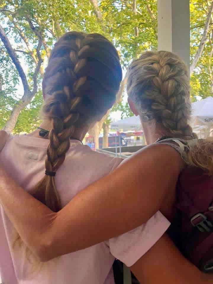 Two pilgrims with long braids, arms around each other, look out at trees and white tents.