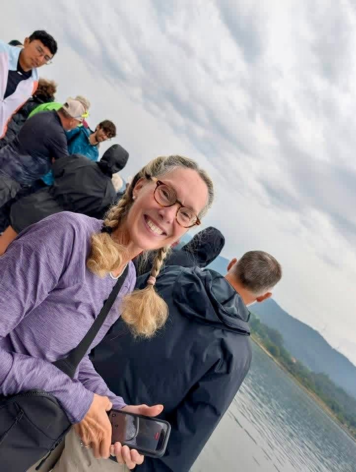 A smiling pilgrim in glasses and a purple shirt takes a selfie on a boat deck, other passengers and a mountain shore behind her.